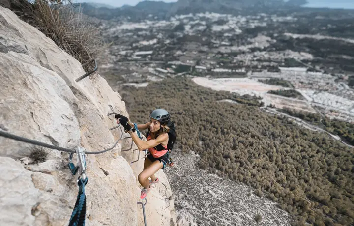 Deportista ascendiendo la vía ferrata del Ponoig en Alicante, con vistas panorámicas hacia Benidorm. Ruta vertical de 160 metros de altura con tramo final en descenso de rapel. Aventura y adrenalina en un entorno natural espectacular.
