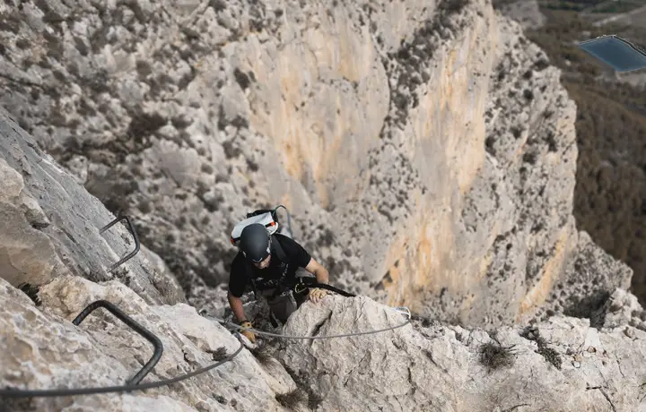 Deportista ascendiendo la vía ferrata del Ponoig en Alicante, con vistas panorámicas hacia Benidorm. Ruta vertical de 160 metros de altura con tramo final en descenso de rapel. Aventura y adrenalina en un entorno natural espectacular.