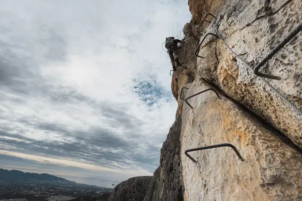 Deportista ascendiendo la vía ferrata del Ponoig en Alicante, con vistas panorámicas hacia Benidorm. Ruta vertical de 160 metros de altura con tramo final en descenso de rapel. Aventura y adrenalina en un entorno natural espectacular.