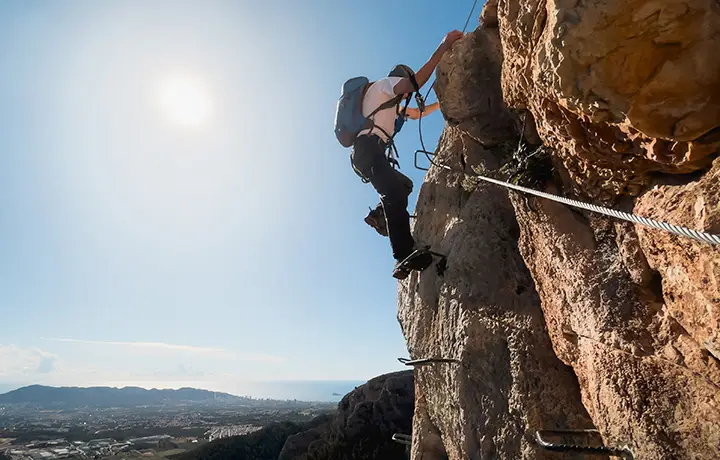 Deportista ascendiendo la vía ferrata del Ponoig en Alicante, con vistas panorámicas hacia Benidorm. Ruta vertical de 160 metros de altura con tramo final en descenso de rapel. Aventura y adrenalina en un entorno natural espectacular.